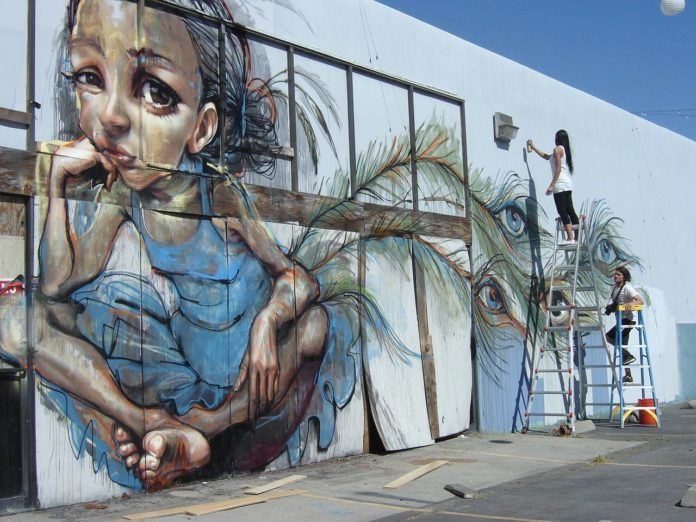 A little girl wearing peacock feathers with eyes looks bored in this enormous street art painting by Graffiti artists Herakut