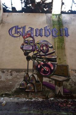 A fed up robot sits beneath the German word for BELIEVE in this street art painting by Pixel Pancho